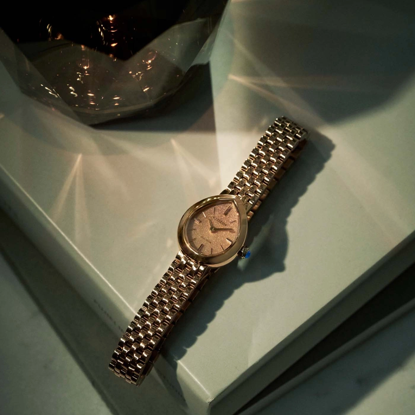 Une montre bracelet dorée en forme de larme sur des livres avec des jeux d'ombre.
