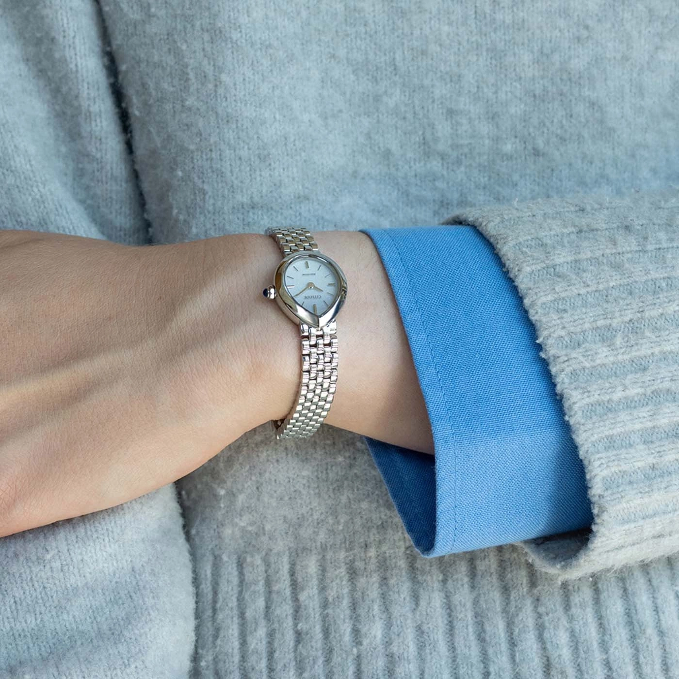 Une main portant une montre argentée sur un pull gris et une chemise bleue.