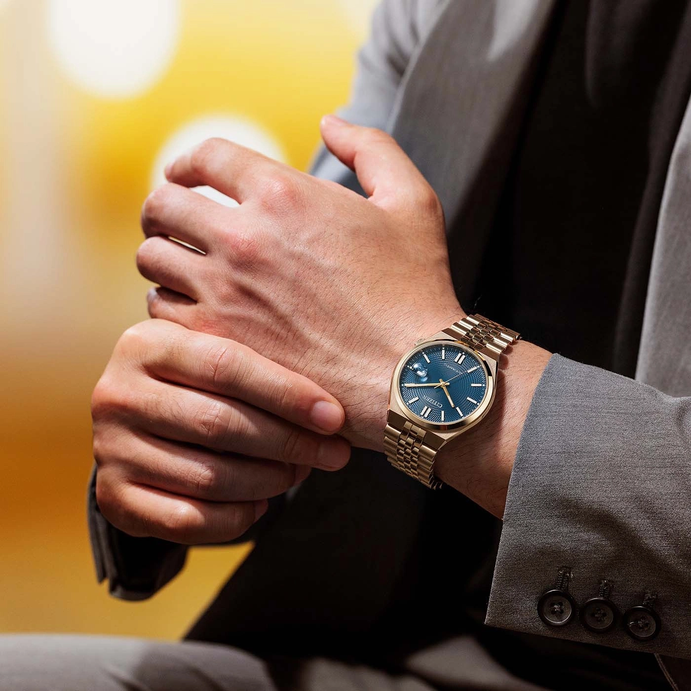 Man in grijs pak stelt zijn gouden horloge met blauwe wijzerplaat bij.