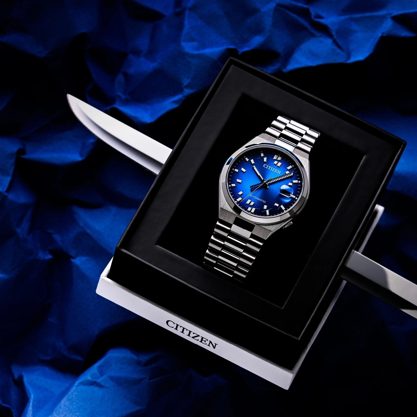 Montre Citizen bleue automatique dans sa boîte, sur fond bleu froissé.