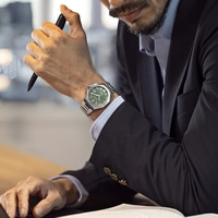 Homme d'affaires avec une montre verte au poignet, tenant un stylo et tapant sur un ordinateur portable.