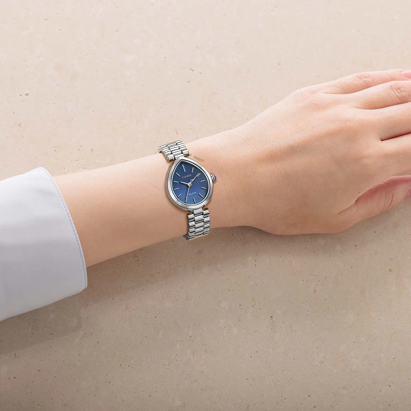 Eine Hand trägt eine blaue Citizen-Uhr mit silbernem Armband auf einem texturierten Hintergrund.