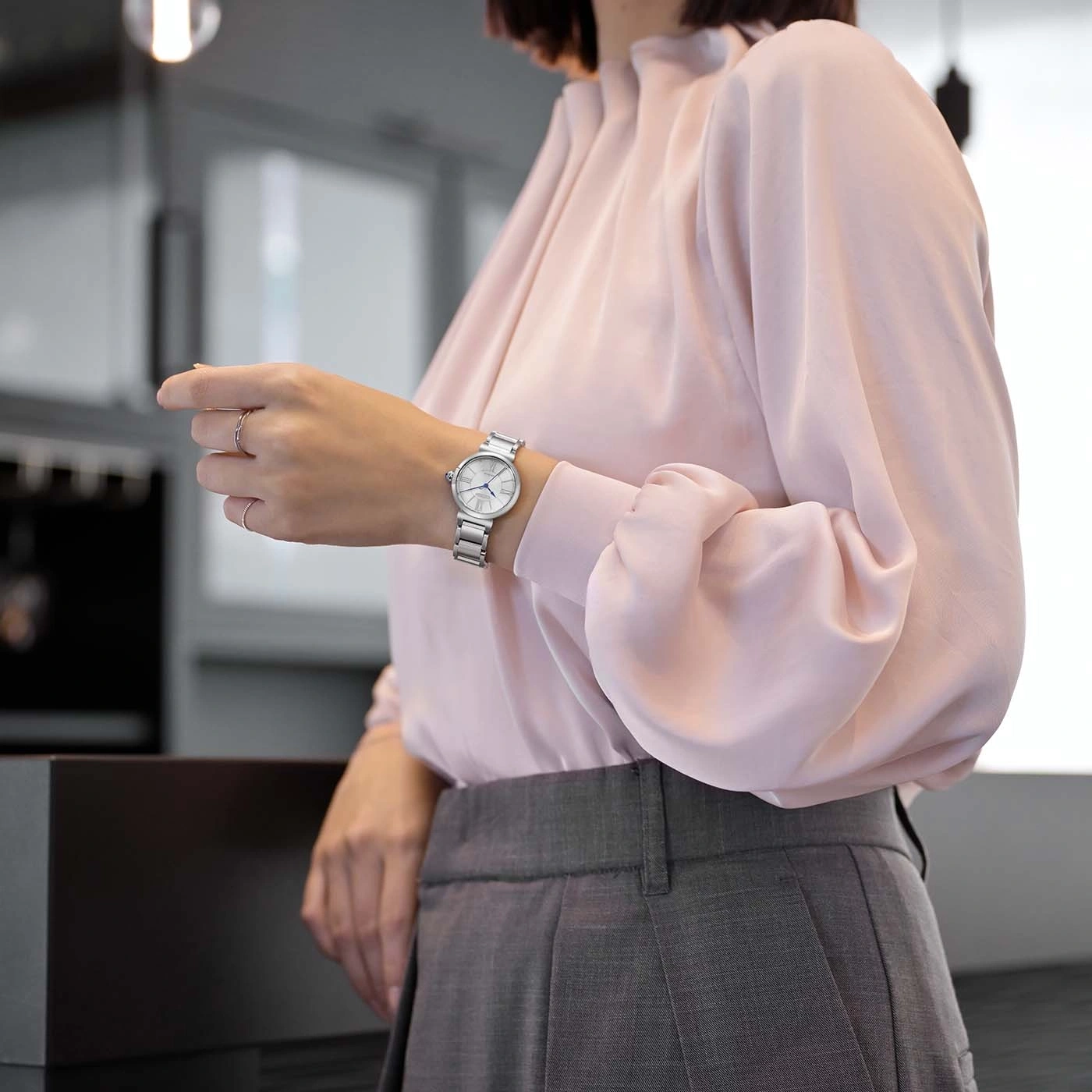 La femme porte une montre argentée à son poignet gauche avec une chemise rose et un pantalon gris.