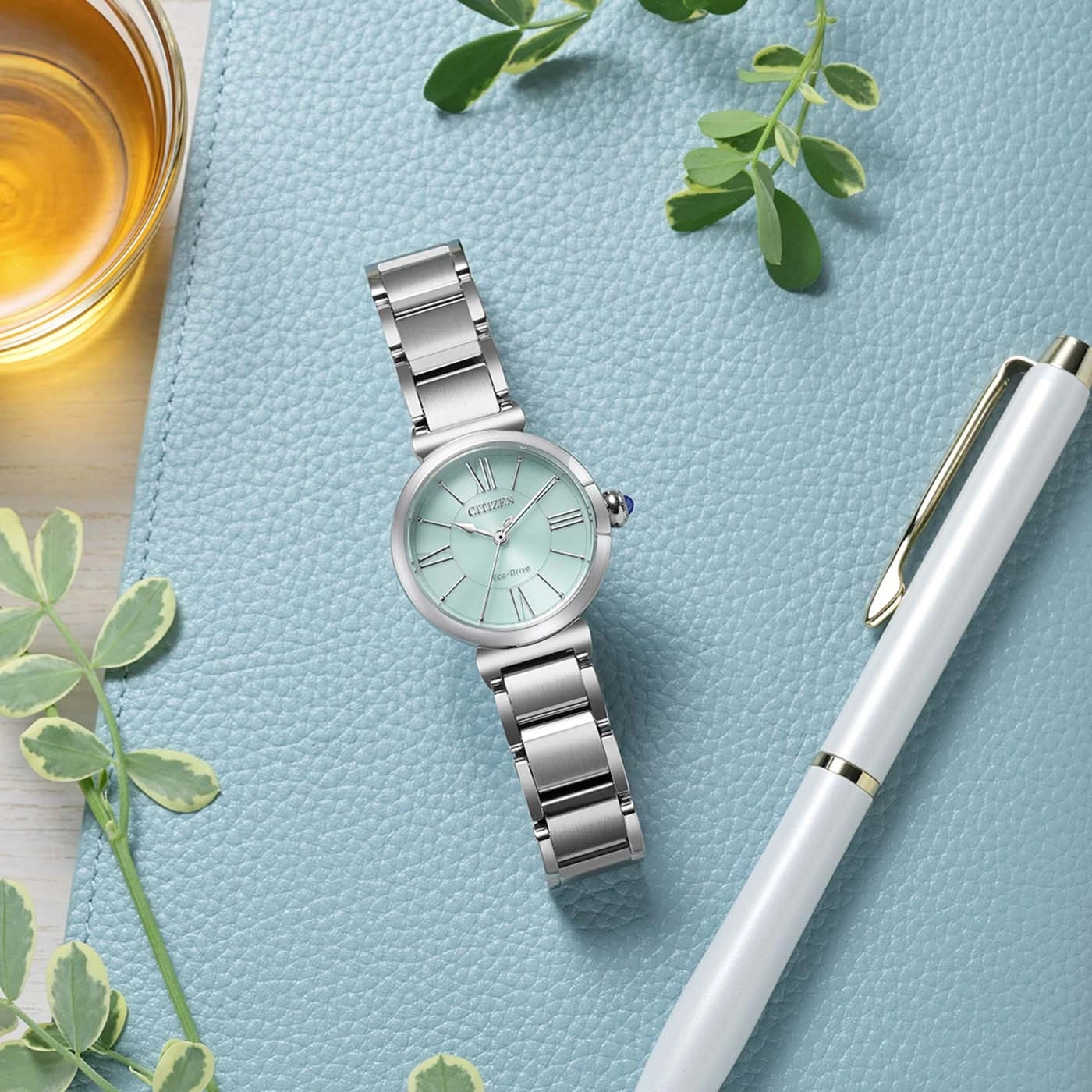 Une montre Citizen Eco-Drive de couleur menthe posée sur un fond bleu sarcelle à côté d'un stylo blanc.