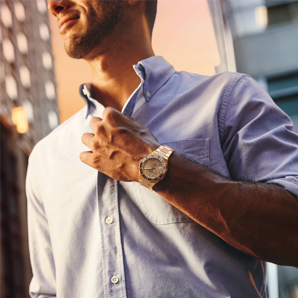 Homme portant une montre en or, ajustant sa chemise, en extérieur, heure dorée, fond de bâtiment.