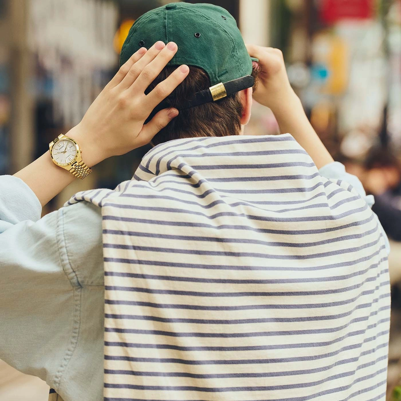 Man met groene pet, gouden horloge en gestreepte trui over zijn schouders.