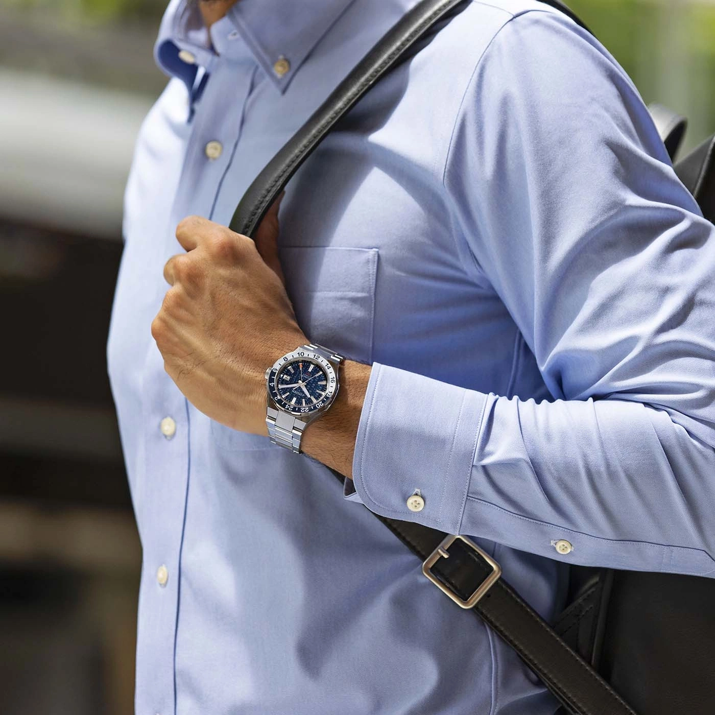 Homme portant une chemise bleue et une montre, sac noir.