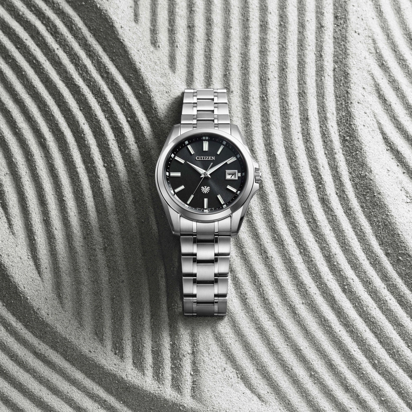 Une montre Citizen argentée repose sur du sable aux rayures parallèles.