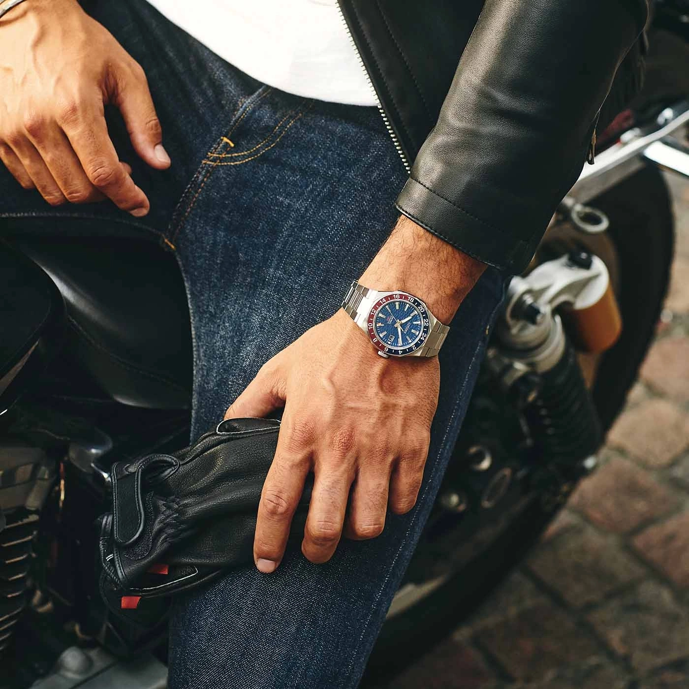 Man wearing a watch, leather jacket, and jeans, holding gloves on a motorcycle.