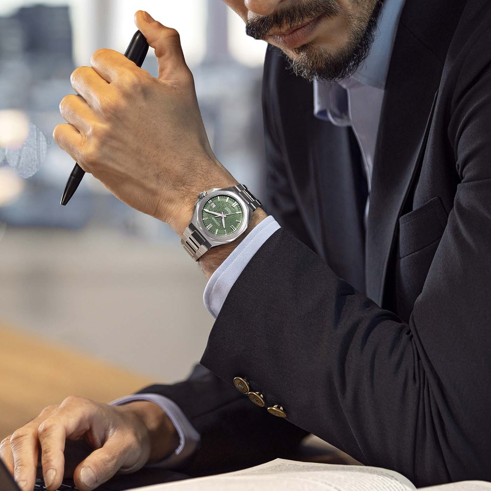 Homme d'affaires avec une montre verte au poignet, tenant un stylo et tapant sur un ordinateur portable.