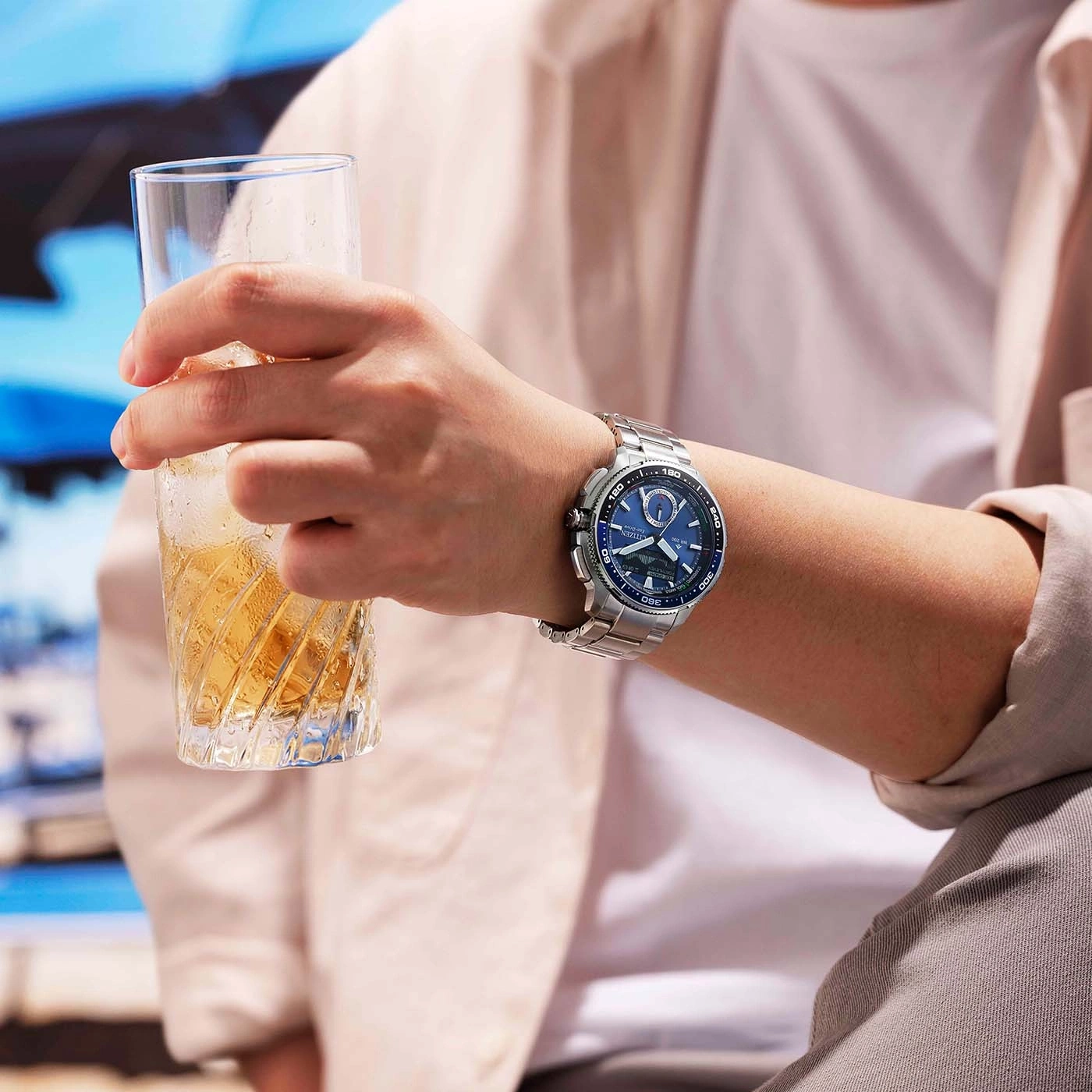 Ein Mann hält ein Glas mit Eiswürfeln und einem bernsteinfarbenen Getränk und trägt eine silberne Armbanduhr.