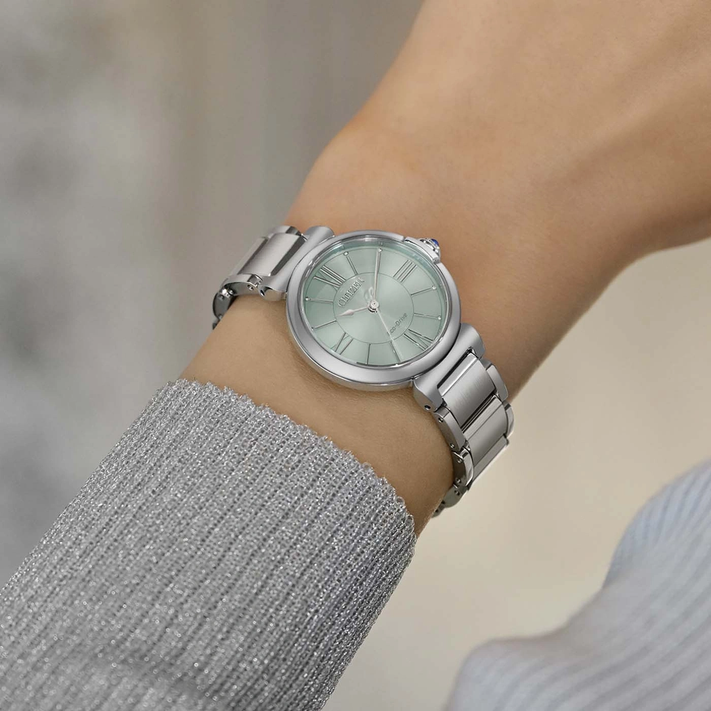 Une femme porte une montre argentée sur son poignet gauche, avec un cadran vert pâle et des chiffres romains.