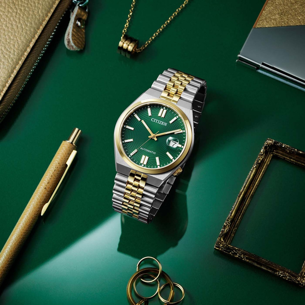 Montre Citizen avec cadran vert, bracelet or et argent, sur surface verte avec accessoires.