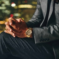 Homme portant une montre en or, mains jointes, costume, lumières de la ville en arrière-plan.